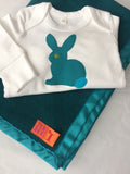 Close up of a white organic cotton baby sleepsuit with long sleeves and scratch mitts and feet. The sleepsuit is folded on top of a teal fleece blanket.  On a clothes hanger against a blue wall. On the front is a hand applique rabbit in fine stripe teal with teal blue tail and lime eye detail. Poppers around the legs to get baby in and out. Available in two sizes 0-3 and 3-6 months, ideal for newborn and premature babies. Handmade in London by Isabee.