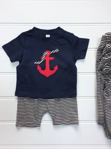 Dark blue cotton T-shirt partially laying on top of a pair of striped cream and navy shorts. T-shirt features an appliquéd red Anchor design, entwined with a striped rope. The clothes lay on a white panneled background. - isabee.co.uk