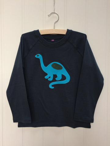Dark blue long-sleeved t-shirt with a turquoise hand-applique dinosaur on the front with grey eye and body detail. This organic cotton top for children is on a hanger with a which background. Made by Isabee in London.
