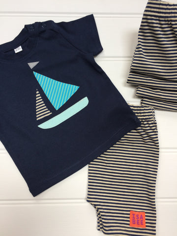 Dark blue cotton T-shirt diagonally laying across of a folded pair of striped cream and navy shorts. Shorts have small orange and purple ISABEE logo. T-shirt features an appliquéd Sailing boat design with a light blue bottom, two stripy sails (one of which matches the shorts, the other in pale greeny-blue shades) and a reflective flag. A small stack of folded shorts sits next to them. The clothes lay on a white panneled background. - isabee.co.uk