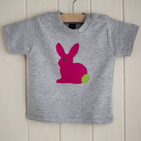 Light grey melange baby's t-shirt featuring a fuchsia pink appliqued rabbit with a leaf green tail. There are two poppers on the t-shirt's shoulder. T-shirt is displayed on a hanger in front of a white panelled background. - isabee.co.uk