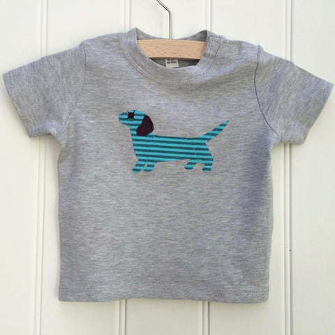 Light grey cotton melange t-shirt for babies with a cheerful applique dachshund sausage dog on the front. The dog is a teal striped with aubergine ear and eye detail. Two poppers on the shoulder for easy wear. T-shirt is displayed on a hanger against a white panelled background. - isabee.co.uk