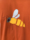 Burnt orange short-sleeved t-shirt for adults with bee applique on the front. Yellow body, white wings, grey head and black eye, antennae and legs. isabee
