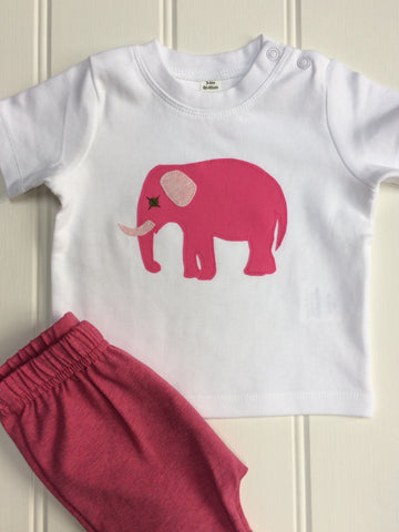 White cotton short-sleeved t-shirt featuring an appliquéd bubblegum pink elephant with soft pink and green accents. There are two subtle poppers (a.k.a. snaps) one shoulder of the top. T-shirt is displayed on an off-white paneled background with a matching pair of pink shorts next to it. - isabee.co.uk