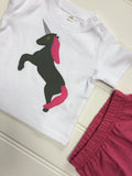 White baby's t-shirt featuring an olive coloured appliquéd Unicorn design with a reflective horn and a bubblegum pink mane and tail. Unicorn is rearing on its hind legs. There are two poppers on the shoulder of the top. Layed on an off-white panneled background beside the top edge of some matching pink shorts. - isabee.co.uk