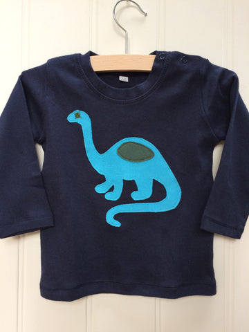 Blue long-sleeve organic cotton t-shirt for babies with turquoise hand-applique dinosaur with grey eye and detail. Two poppers on the shoulder for easy wear. The top is on a hanger with a white background. Hand made in the Isabee London studio.