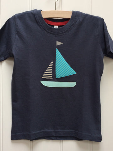 Dark blue cotton T-shirt featuring an appliquéd Sailing boat design with a light blue bottom, two stripy sails (one of which matches the shorts, the other in pale greeny-blue shades) and a reflective flag. The t-shirt is mounted on a hanger against a white panneled background. - isabee.co.uk