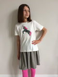 Young girl wearing a cream coloured cotton children's t-shirt featuring an olive coloured appliquéd Unicorn design with a reflective horn and a bubblegum pink mane and tail. Unicorn is rearing on its hind legs. Girl wears matching grey skirt and pink leggings and stands, with one hand on her hip, in front of a white wall. - isabee.co.uk