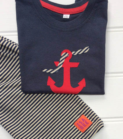 Folded dark blue cotton T-shirt partially laying across of a folded pair of striped cream and navy shorts. Shorts have small orange and purple ISABEE logo. T-shirt features an appliquéd red Anchor design, entwined with a striped rope. The clothes lay on a white panelled background. - isabee.co.uk