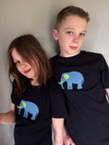 Girl and boy wearing Black cotton t-shirts featuring an appliquéd, meadow blue elephant with green and red accents. The two children lean against each other smiling at the camera. - isabee.co.uk