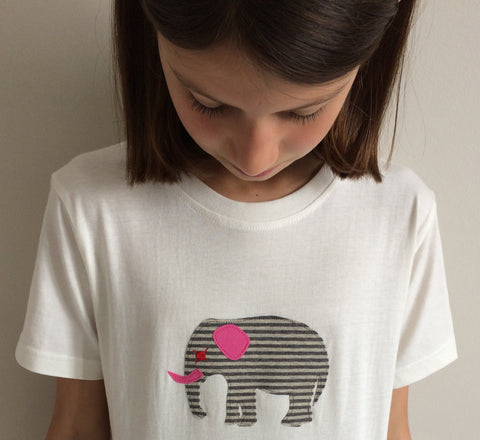 Girl looking down at the t-shirt she is wearing. Cream coloured t-shirt featuring an appliquéd, grey striped elephant with pink and red accents. Off-white wall behind model. - isabee.co.uk