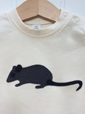 Close up photo of a natural white organic cotton, short-sleeved t-shirt on a hanger with a little applique mouse on the front. The mouse made of fine stripe grey cotton melange, with black eye, ears and tail. Designed and hand made in the Isabee London studio. Available in four sizes for babies (3 -24 months). Includes Isabee easy-wear neck with poppers. Also available for ages 2 to 5 years.