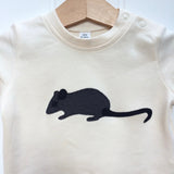 Close-up photo of a natural white organic cotton, short-sleeved t-shirt on a hanger with a little applique mouse on the front. The mouse made of fine stripe grey cotton melange, with black eye, ears and tail. Designed and hand made in the Isabee London studio. Available in four sizes for babies (3 -24 months). Includes Isabee easy-wear neck with poppers. Also available for ages 2 to 5 years.