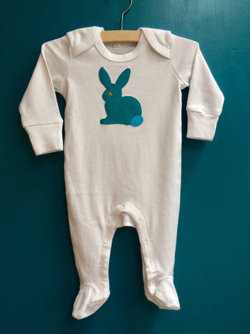 White organic cotton baby sleepsuit with long sleeves and scratch mitts and feet. On a clothes hanger against a blue wall. A hand applique rabbit in fine stripe teal with teal blue tail and lime eye detail is on the front. Poppers around the legs to get baby in and out. In sizes for newborn and premature babies. Handmade in London -isabee.co.uk