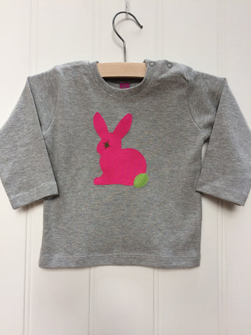 Baby Rabbit long-sleeved t-shirt on a hanger.  The top is a soft grey colour and  features an applique rabbit in fuschia pink with a leaf green tail and eye detail. Shoulder poppers on one side for easy wear. Hand made by Isabee in London.