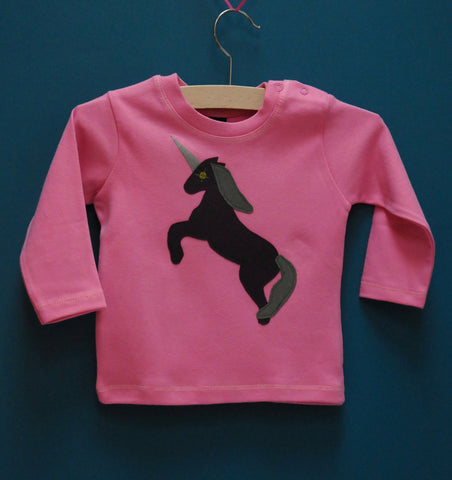 Bubblegum pink baby's long sleeved t-shirt featuring an augergine coloured Appliquéd Unicorn design with a reflective horn and a olive green mane and tail. Unicorn is rearing on its hind legs. There are two poppers on the shoulder of the top. T-shirt is on a hanger in front of a teal background. - isabee.co.uk