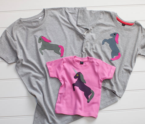 Three t-shirts with hand applique unicorn design on the front, displayed on white background. The two adult and child t-shirts are grey melange and feature a darker grey unicorn rearing on its hind legs with a reflective horn and pink mane and tail. The baby t-shirt is bubblegum pink with an aubergine coloured unicorn and olive green mane and tail. - isabee.co.uk