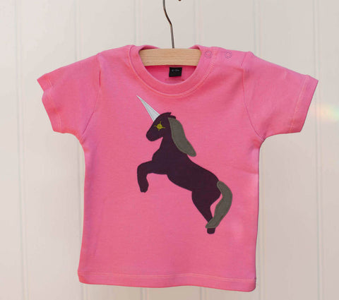 Bubblegum pink baby's t-shirt featuring an augergine coloured appliquéd Unicorn design with a reflective horn and an olive green mane and tail. Unicorn is rearing on its hind legs. There are two poppers on the shoulder of the top. T-shirt is on a hanger in front of an off-white background. - isabee.co.uk