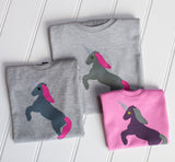 Three folded t-shirts on white panelled background each has a hand applique unicorn design on the front. The two adult and child t-shirts are grey melange with a darker grey unicorn rearing on its hind legs with a reflective horn and pink mane and tail. The baby t-shirt is bubblegum pink with an aubergine coloured unicorn and olive green mane and tail. - isabee.co.uk