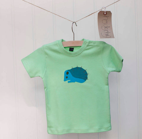 Pale mint green coloured short-sleeved t-shirt for babies aged 3-24 months - on a hanger. It has a hand-sewn applique hedgehog on the front. The hedgehog is pale and dark blue with black detail with hand-sewn eye, ear and spikes. Two poppers on the neck to help get baby in and out. Made in London by Isabee - ethically sourced cotton.